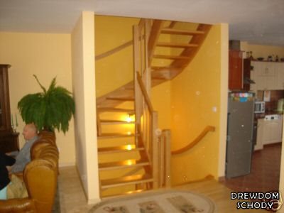 Jasne drewniane schody kręcone z poręczą, widoczne z salonu w domu w Krępsku, oświetlone od dołu.